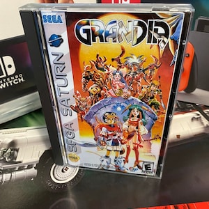 May include: A Sega Saturn video game case with a red, orange, and yellow background. The game is called "Grandia" and features a group of characters in a fantasy setting. The case is in good condition and has a clear plastic cover.