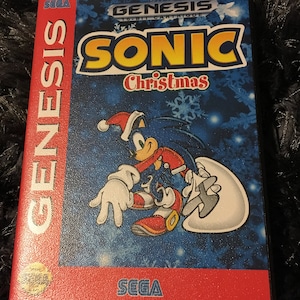 May include: A Sega Genesis game cartridge for "Sonic Christmas." The game case features a blue background with snowflakes and Sonic the Hedgehog wearing a Santa hat. The Sega and Genesis logos are visible.