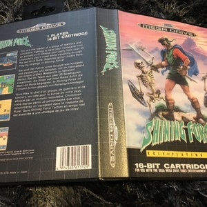 May include: A Sega Mega Drive game cartridge for Shining Force, a role-playing game. The cartridge is black with a silver label that says "Shining Force" and "16-bit Cartridge". The back of the cartridge has a description of the game in English, French, and German.