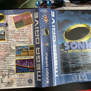 May include: A SEGA Mega Drive game case for "Sonic and the Mystic Gems." The blue case features the game title and a gold ring graphic over water. The spine reads "MEGA DRIVE." The back displays screenshots and game information.