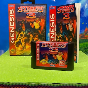 May include: A Sega Genesis video game cartridge for Streets of Rage 3. The cartridge is black with a blue label that says "Streets of Rage 3" and has a picture of the game's characters. The cartridge is sitting on a green surface.