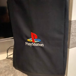 Może przedstawiać: Czarny pokrowiec z tkaniny z haftowanym logo PlayStation w kolorze czerwonym, niebieskim, żółtym i białym.