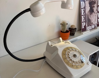 Pode incluir: Um telefone de disco branco reaproveitado como candeeiro de secretária. A base apresenta um mostrador circular bege com números e letras. Um braço flexível preto tipo pescoço de ganso estende-se da base, suportando o auscultador como luz.