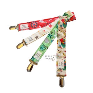 May include: Four fabric pacifier clips with metal clasps are displayed against a white background. The clips feature various patterns, including nautical, stars, and cartoon illustrations. Each clip has a small label that reads "Little Arth Studio."