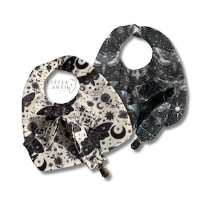 May include: Two baby bibs featuring moth and floral designs. One is off-white with black moths, flowers, and moons. The other is black with a celestial moth pattern. Both have a pocket and a clip for a soother. The text "LITTLE ARTH STUDIO" is visible.