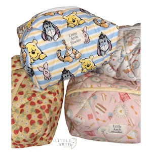 May include: A stack of quilted cosmetic bags with various patterns. One bag features Winnie the Pooh characters on a blue and yellow striped background. Other bags display strawberry, bicycle, and cake designs. The bags have a zipper closure and the text "Little Arth Studio".