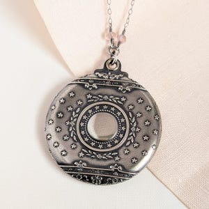 May include: A silver-toned pendant necklace with a round, ornate locket design. The locket features a central circle, surrounded by a ring of stars and floral details. The pendant hangs from a delicate chain with pink beads.