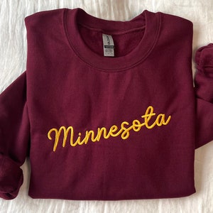 Puede incluir: Sudadera color burdeos con la palabra "Minnesota" bordada en oro en cursiva en la parte delantera.