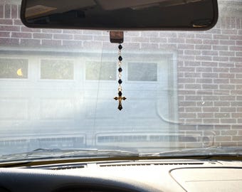 May include: A car rearview mirror ornament featuring a vertical chain of small, faceted black beads connected by silver-toned metal links, ending in a gold-toned crucifix pendant with a central figure, hanging from the center of the mirror.