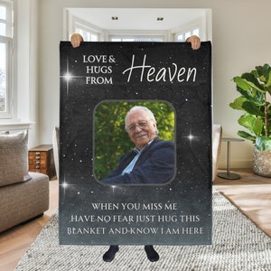 以下が含まれることがあります： 「Love & Hugs From Heaven」と書かれた白い文字が入った黒い毛布。正方形のフレームに入った男性の写真が特徴です。毛布には「When you miss me, have no fear just hug this blanket and know I am here」というテキストも入っています。