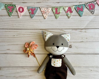 Puede incluir: Un juguete de peluche de gato gris que lleva un mono marrón con lunares blancos y dorados. El juguete de gato sostiene un pequeño molinillo de viento naranja y blanco.