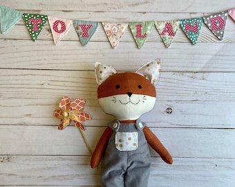 May include: A plush fox toy wearing grey overalls with white and gold polka dots. The fox has orange fur, a white face, and black eyes. The toy is holding a yellow and orange pinwheel.