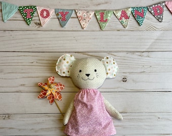 May include: A plush toy mouse wearing a pink dress with white polka dots and gold glitter ears. The mouse is holding a colorful pinwheel. The background is a white wooden surface with a banner that reads "TOYLAND" in pink letters.