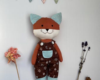 May include: A handmade fox doll with brown and blue accents. The fox wears brown overalls with a floral pattern and a light blue pocket. The doll has a white face, brown ears, and a smiling expression. A banner that says "TOYLAND" hangs above the doll.