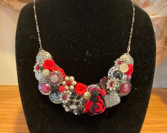 May include: A statement necklace featuring a cluster of red and black fabric roses, rhinestones, and vintage buttons. The necklace is on a silver chain.