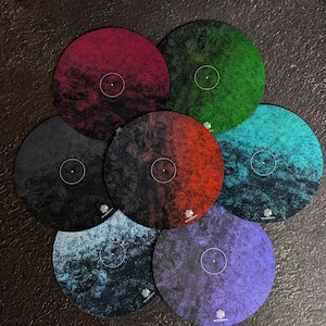 以下が含まれることがあります： 赤、緑、青、紫など、さまざまな色の7つの円形コースターのコレクション。各コースターは、テクスチャ加工された表面、中央の白い点、小さなロゴが特徴です。コースターは円形に配置されています。