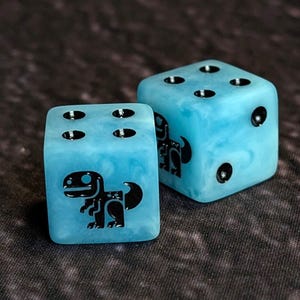 Puede incluir: Dos dados de seis caras con acabado en resina veteada de color azul claro. Cada dado presenta puntos negros y un gráfico negro de un dinosaurio estilizado en una cara. Los dados tienen esquinas redondeadas y una superficie lisa y pulida.