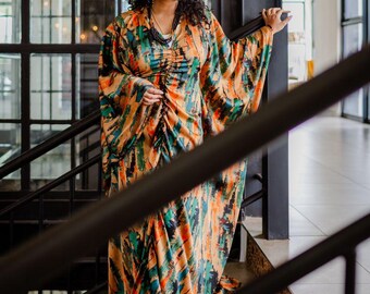 May include: A woman wearing a long, flowing orange, green and blue patterned dress with a gathered waist. She is standing on a staircase with a black railing.