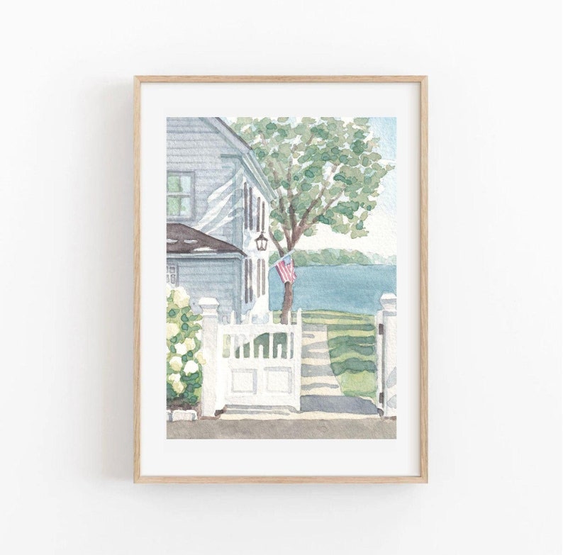 May include: Watercolor painting of a coastal house with a white picket fence and gate. The house is light blue with a dark roof, and an American flag hangs from a tree. The artwork is framed in a light wood frame.