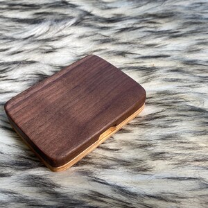 May include: A rectangular wooden box with a dark brown top and a lighter wood base. The box is closed and sits on a textured, white and gray surface. The box appears to be made of two different types of wood.