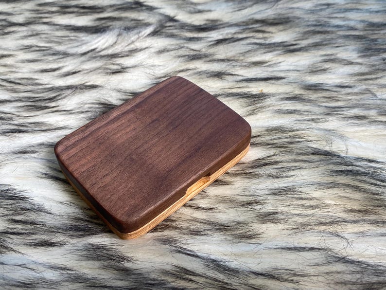 May include: A rectangular wooden box with a dark brown top and a lighter wood base. The box is closed and sits on a textured, white and gray surface. The box appears to be made of two different types of wood.