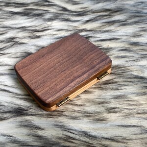 May include: A rectangular wooden box with a dark brown top and a lighter wood base. The box is closed with small brass hinges. The wood grain is visible on the top surface.