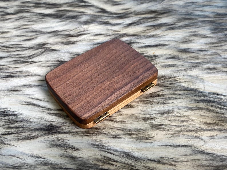 May include: A rectangular wooden box with a dark brown top and a lighter wood base. The box is closed with small brass hinges. The wood grain is visible on the top surface.