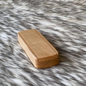 May include: A small, rectangular wooden box with a lid. The box is made of light-colored wood and has a simple, minimalist design. The box is closed and appears to be empty. The box is sitting on a textured, faux fur surface.