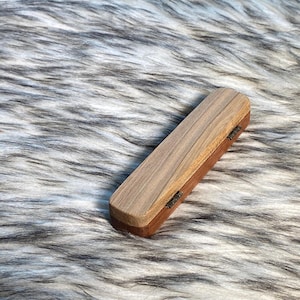 May include: A rectangular wooden box with a light brown lid and a darker brown base. The box is closed with two small metal hinges. The box is resting on a fluffy, gray and white surface.