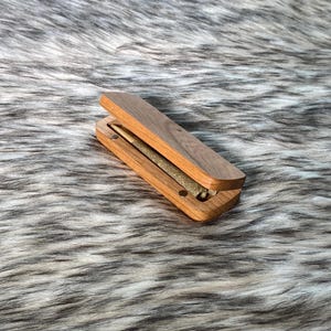 May include: A wooden box with a hinged lid, open to reveal a rolled item. The box is made of a light brown wood with a darker wood lid. The item inside is a rolled cylinder, likely a cigarette or similar product.