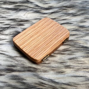 May include: A small, rectangular wooden box with a hinged lid. The box is made of light-colored wood with a visible grain pattern. The lid is closed, and the box appears to be empty. The box is resting on a fluffy, faux fur surface.