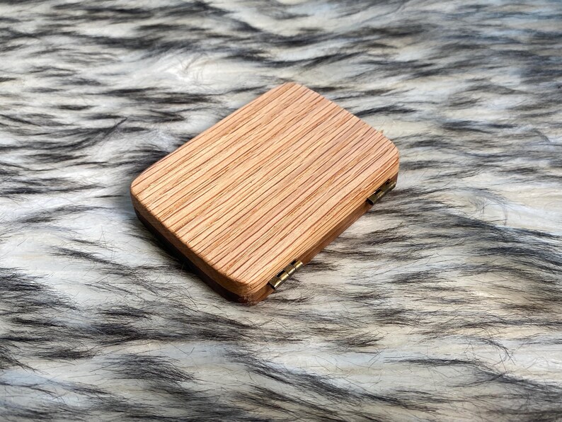 May include: A small, rectangular wooden box with a hinged lid. The box is made of light-colored wood with a visible grain pattern. The lid is closed, and the box appears to be empty. The box is resting on a fluffy, faux fur surface.