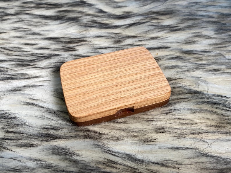 May include: A rectangular wooden box with a light brown, striped top and a darker brown base. The box is closed and sits on a fluffy, faux fur surface with black and white streaks. The wood grain is visible.