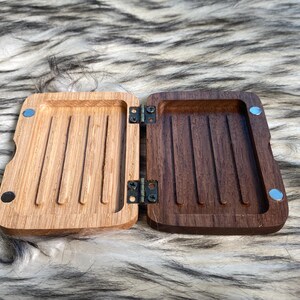 May include: An open, hinged wooden box with a light brown interior on one side and a dark brown interior on the other. The box has a rectangular shape with rounded edges and is held together by hinges and small magnets.
