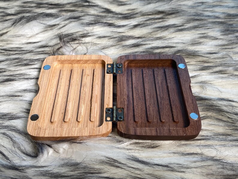 May include: An open, hinged wooden box with a light brown interior on one side and a dark brown interior on the other. The box has a rectangular shape with rounded edges and is held together by hinges and small magnets.