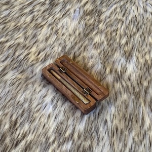 May include: An open, rectangular wooden box with a hinged lid, revealing two cylindrical objects inside. The box and contents are set against a backdrop of a fluffy, textured, light brown and white surface.