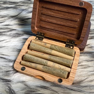 May include: A wooden box with a hinged lid, open to reveal several rolled cigarettes. The box is made of a light-colored wood, with a darker wood lid. The cigarettes are wrapped in tan paper. The box is resting on a fluffy, gray and white surface.