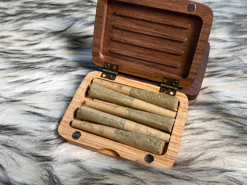 May include: A wooden box with a hinged lid, open to reveal several rolled cigarettes. The box is made of a light-colored wood, with a darker wood lid. The cigarettes are wrapped in tan paper. The box is resting on a fluffy, gray and white surface.