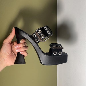 Può includere: Tacchi alti neri con plateau, tacco spesso e due fibbie argentate. I tacchi hanno più occhielli argentati.