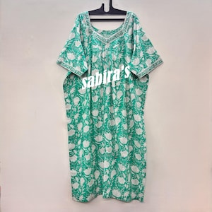 May include: A green and white floral print dress with short sleeves and a square neckline. The dress is hanging on a black hanger. The text "sabira's" is printed on the front of the dress.