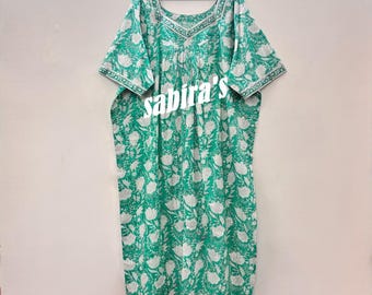 May include: A green and white floral print dress with short sleeves and a square neckline. The dress is hanging on a black hanger. The text "sabira's" is printed on the front of the dress.