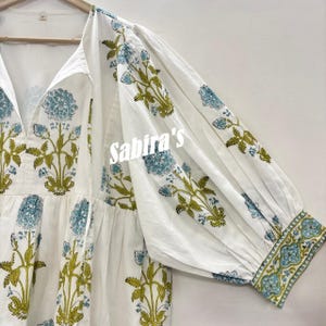 Pu&ograve; includere: Abito in cotone bianco con scollo a V e maniche lunghe. L'abito presenta un motivo floreale nei toni del blu e del verde. Il testo "Sabira's" &egrave; visibile sulla parte anteriore dell'abito.