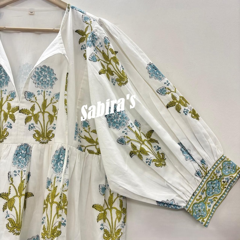 Pu&ograve; includere: Abito in cotone bianco con scollo a V e maniche lunghe. L'abito presenta un motivo floreale nei toni del blu e del verde. Il testo "Sabira's" &egrave; visibile sulla parte anteriore dell'abito.