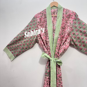 May include: A robe featuring a floral pattern in shades of pink and sage green, with a white text overlay reading Sabira's. The garment includes a light green and white striped trim along the collar, cuffs, and a matching tie belt at the waist.