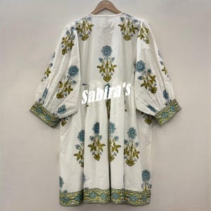 Pu&ograve; includere: Abito bianco con stampa floreale blu e verde. L'abito ha uno scollo a V, maniche lunghe e vita arricciata. Le maniche e l'orlo sono rifiniti con un bordo a fantasia. Il testo "Sabira's" &egrave; visibile sul davanti.