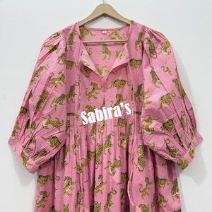 May include: A light pink dress featuring a repeating pattern of yellow and black tigers. The dress has a V-neckline with tie closures, three-quarter length puff sleeves with cuffs, and a gathered waist. The text Sabira's is printed in white.