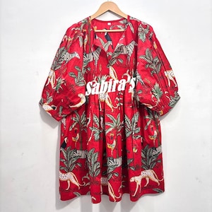 May include: A red dress featuring a vibrant jungle print with zebras, leopards, and tropical birds among palm trees. The garment has a V-neckline, gathered waist, and voluminous three-quarter length sleeves. The text "sabira's" is printed in white.