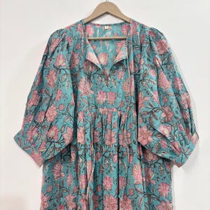 Może przedstawiać: Turkusowa sukienka z kwiatowym wzorem w kolorze różowym i brązowym. Sukienka ma dekolt w kształcie litery V, długie rękawy i marszczoną talię. Tkanina wydaje się lekka, odpowiednia na ciepłą pogodę.