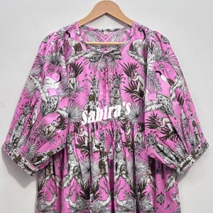 Könnte beinhalten: Eine rosa Bluse mit einem schwarz-weißen Dschungel-Print, der Affen, Leoparden und tropisches Blattwerk zeigt. Das Kleidungsstück hat dreiviertellange Puffärmel, einen Rundhalsausschnitt mit Bindeband und den in Weiß gedruckten Text Sabira's.