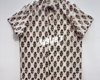 May include: A short-sleeve button-down shirt in an off-white color featuring a repeating floral pattern in dark red and black. The shirt has a standard collar, a front button placket, and a chest pocket. The text Sabira's is printed in white.